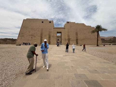       Travelers walk toward the massive pylons of an ancient sandstone temple under streaked clouds.
  