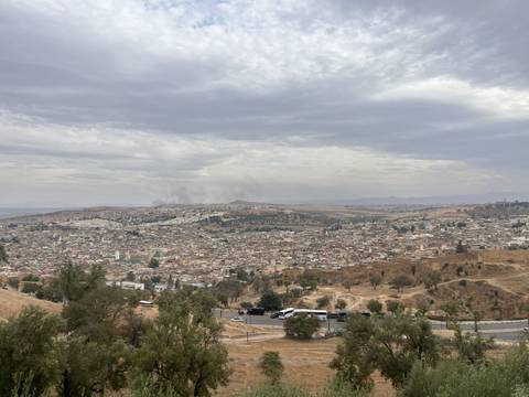       Wide, hazy panorama of Fes spreads beneath an overcast sky, with smoke plumes rising in the distance.
  