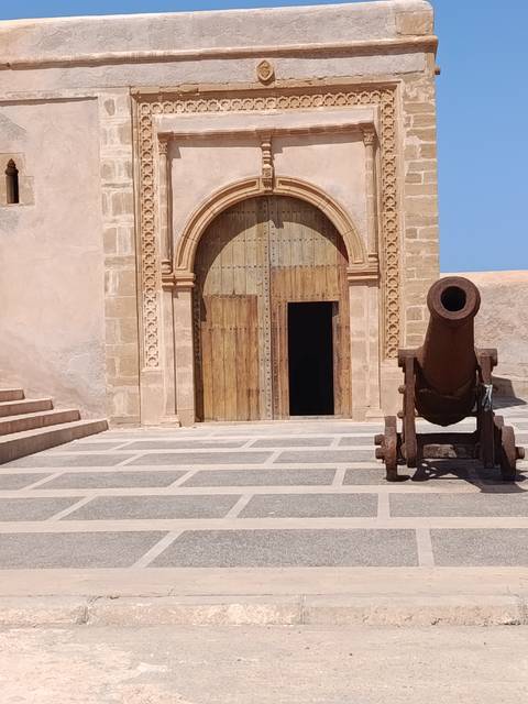       Old rusted cannon facing a carved wooden doorway in a stone courtyard.
  