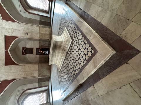       Stone cenotaph inside a Mughal mausoleum surrounded by patterned marble flooring.
  