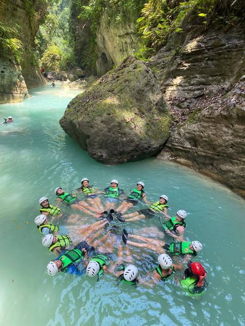       Adventurers wearing helmets and life vests float in a circle in turquoise canyon waters beneath a large boulder.
  