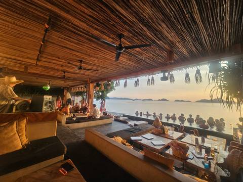       Lively infinity-pool bar at sunset where guests relax with sea views and DJ music under a bamboo roof.
  