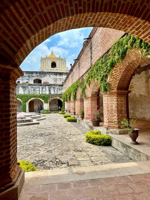       Peaceful cloister courtyard with brick arches, ivy, and potted plants inside an Antigua convent.
  
