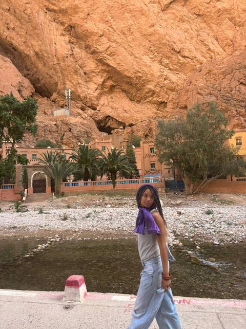       A young girl in a purple scarf poses with palm trees and orange rock walls of Todra Gorge behind her.
  