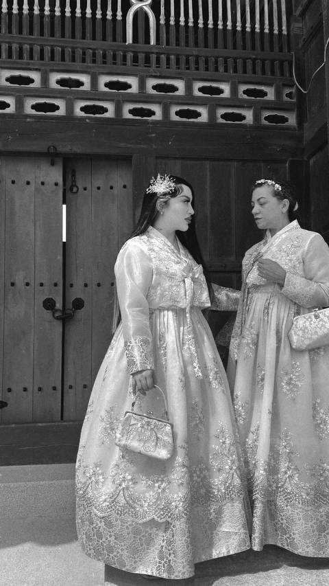       Black-and-white close-up of two women wearing traditional hanbok dresses conversing at a wooden gate.
  