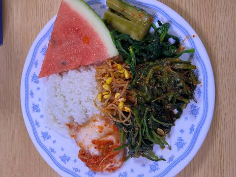       Close-up plate with rice, kimchi, greens, bean sprouts and a slice of watermelon on a wooden table.
  