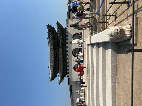       Wide view of Gwanghwamun Gate with visitors on steps and courtyard under clear blue sky.
  