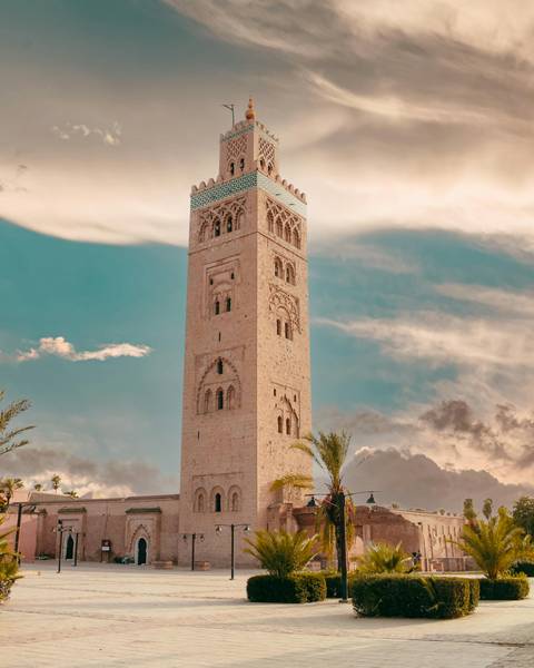       Tall sandstone minaret with intricate carvings rises against a turquoise sky scattered with clouds and framed by a palm frond.
  