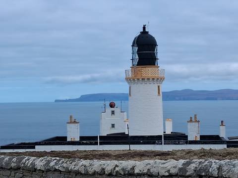       White lighthouse with black lantern rises above the calm blue sea on a grey coastal day in northern Scotland.
  