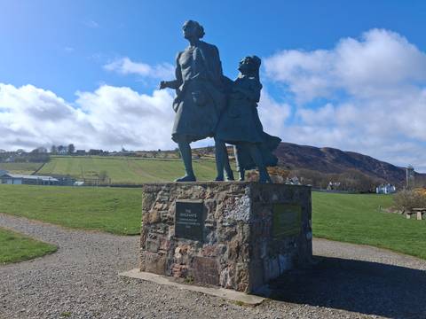       Stone monument of two Highlanders commemorates the emigrants against a backdrop of rolling fields and blue sky.
  