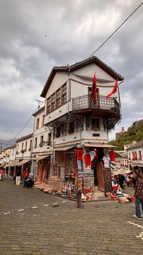       Historic Ottoman-era building with wrought-iron balcony and stalls selling souvenirs and flags.
  