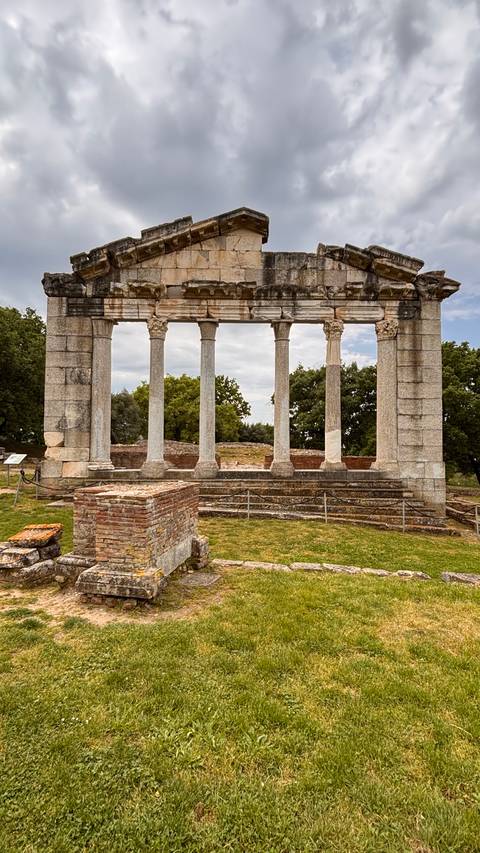       Ancient marble colonnade of a ruined temple set against grassy field and cloudy sky.
  
