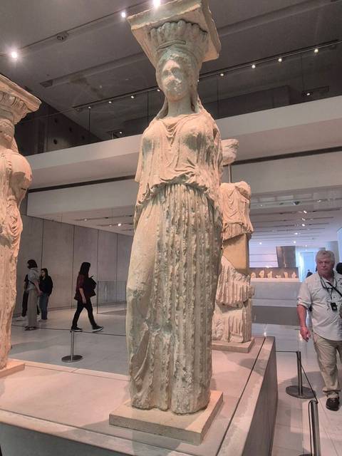       Marble Kore statue displayed inside a modern museum hall with visitors observing ancient artifacts.
  