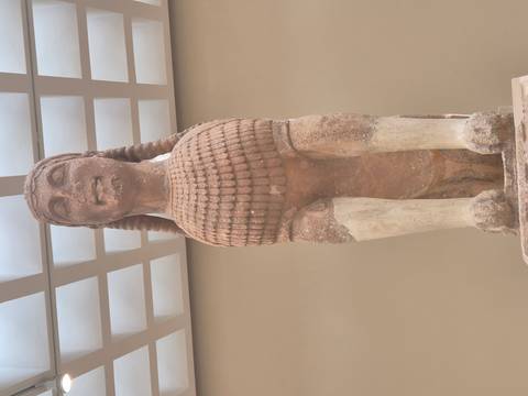       Ancient Greek sphinx statue with detailed scaled chest displayed under a skylight in a museum.
  