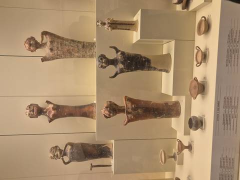       Display of small terracotta figurines and pottery vessels arranged on museum shelves with descriptive labels.
  