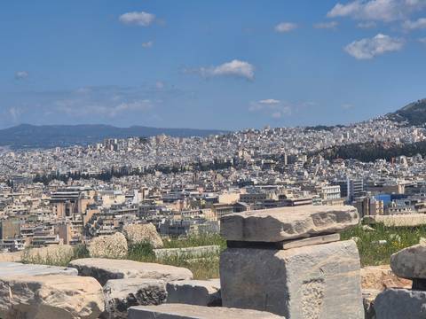       Panoramic view of Athens' densely packed buildings sprawling across hills beneath a blue sky.
  