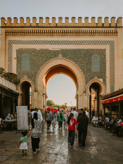       Crowds pass through the ornate green-tiled Bab Bou Jeloud gate glowing in late afternoon light.
  