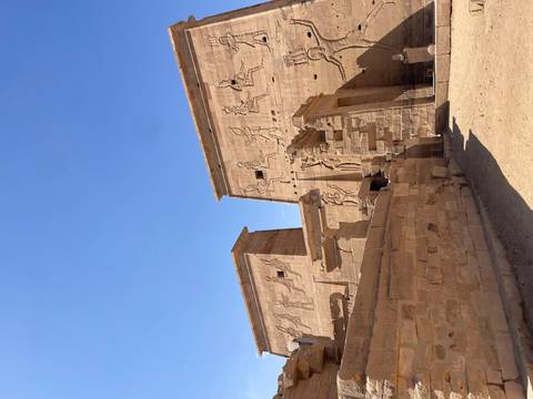       Carved reliefs of figures stride across tall sandstone walls at Philae Temple under a bright sky.
  