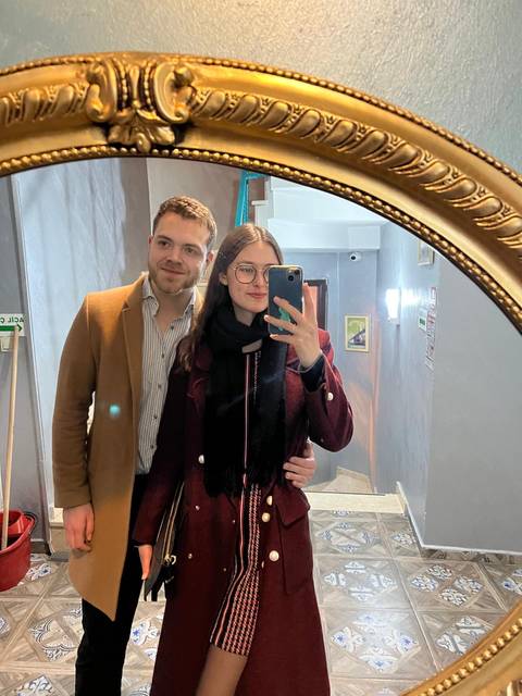       A young couple takes a mirror selfie indoors wearing winter coats and scarves.
  