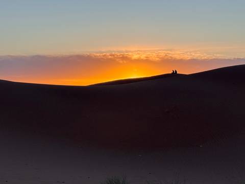       Silhouettes on a dune watch the sun set behind rolling Sahara sand waves.
  