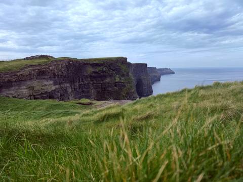       Iconic Cliffs of Moher rising sheer above the Atlantic with grassy foreground under mottled sky.
  