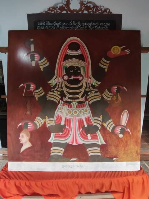      Traditional Sri Lankan mural depicting a multi-armed deity on a red background inside a temple.
  