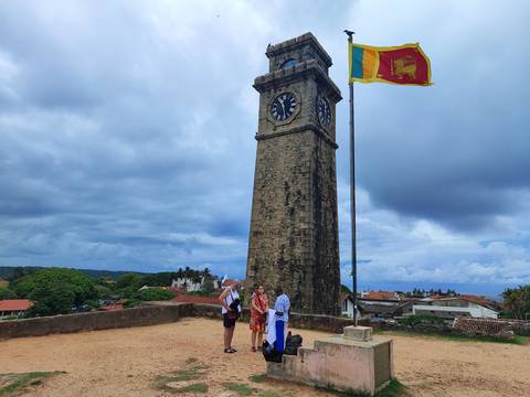       Historic stone clock tower at Galle Fort with Sri Lankan flag and tourists posing below on a cloudy day.
  