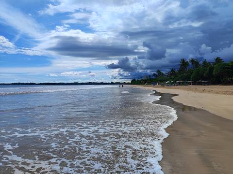       Long stretch of shoreline with gentle surf and dramatic cloud layers over the sea.
  