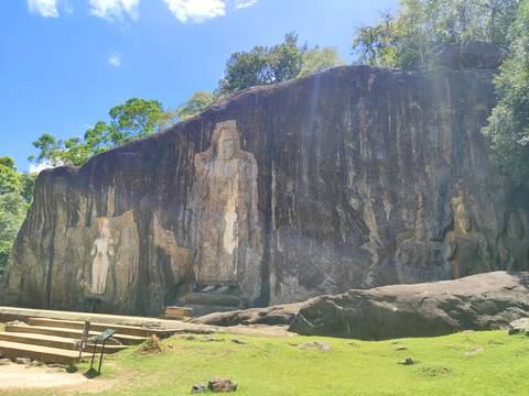       Huge cliff face bearing ancient standing Buddha reliefs carved into dark rock under sunlight.
  