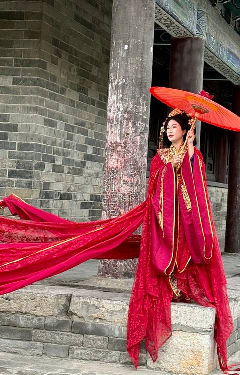       A woman in an ornate red traditional Chinese robe poses gracefully beside an aged stone wall, holding a parasol with a flowing train of fabric.
  
