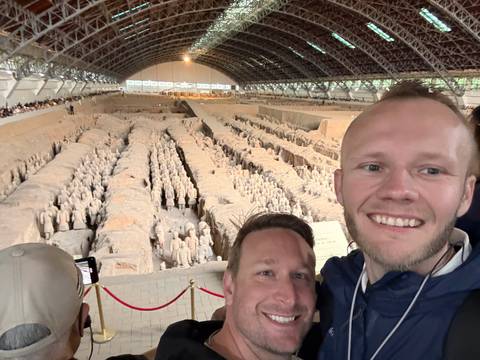       Two smiling travelers take a selfie overlooking the vast Terracotta Army excavation hall filled with warrior statues.
  