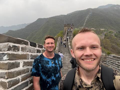       Two friends capture a selfie while walking along a stone section of the Great Wall of China winding over green mountains.
  