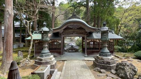       Ornate wooden temple gate flanked by stone lanterns amid tall cedar trees.
  