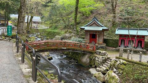       Small red wooden bridge crossing a clear stream beside moss-covered shrines in a forest setting.
  