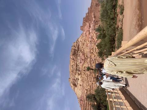       Visitors cross a bridge toward the clay hillside village of Aït Benhaddou on a sunny day.
  
