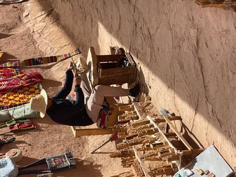       Local artisan in straw hat carving wooden souvenirs in a narrow earthen alleyway.
  