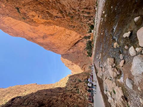       Towering red cliffs of Todra Gorge with a shallow stream and visitors walking along the pathway.
  