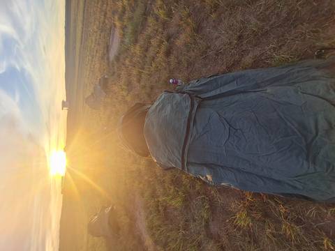      A rolled-out swag lies on grassy ground facing a bright sunrise on an open plain.
  
