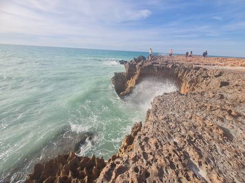       Waves crash into jagged coastal rock formations while a few visitors walk along the cliff top.
  