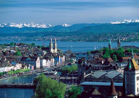       Aerial view of Zurich’s Old Town, lake and distant snow-capped Alps under blue skies.
  