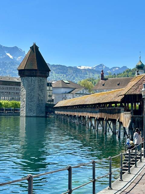       Lucerne’s wooden Chapel Bridge spans calm blue water with alpine peaks behind.
  