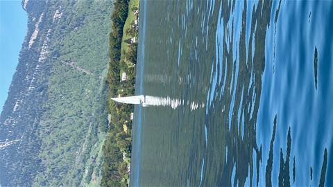       A single sailboat glides across a calm mountain lake reflecting green hills.
  