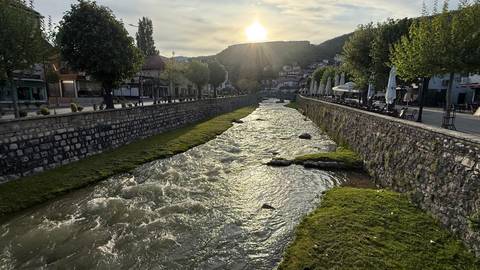       Fast-flowing river flanked by stone embankments and tree-lined walks as the sun sets behind a hilltop town.
  