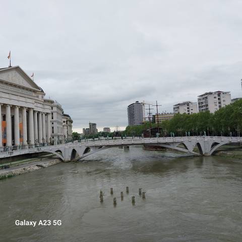       Elegant white pedestrian bridge and riverside museums in a modern Balkan city under cloudy skies.
  