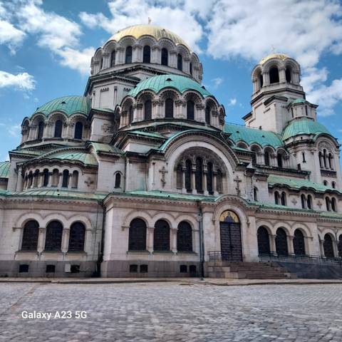       Grand Orthodox cathedral with green copper domes, ornate arches and bright blue sky.
  