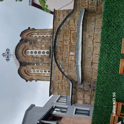       Small stone Orthodox chapel with circular windows and brick detailing beneath a cloudy sky.
  