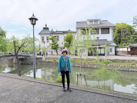       Traveler poses beside a willow-lined canal and historic white buildings in a quaint Japanese town.
  