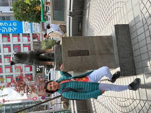       Visitor smiles next to the famous Hachikō dog statue outside a busy Shibuya station square.
  