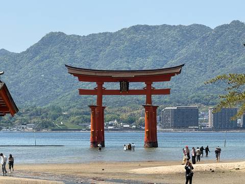       Iconic red torii gate of Itsukushima Shrine rises from the sea with mountains and visitors wading nearby.
  