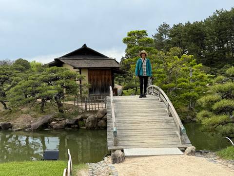       Traveler stands on a small arched wooden bridge in a manicured Japanese garden.
  
